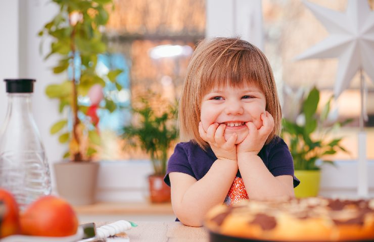 Bambina seduta a tavola con frangetta che ride con il volto appoggiato sulle mani