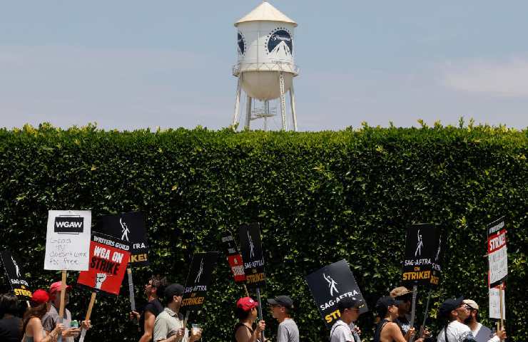 Davanti ad un'alta siepe con dietro il logo di Paramount Pictures ci sono dei manifestanti che scioperano sotto il sole con dei cartelli