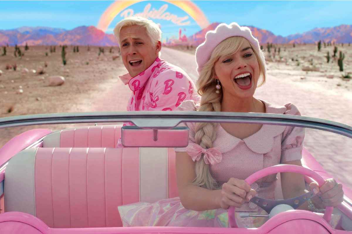 Berbie e Ken nella loro macchina rosa per una strada americana con dietro un arcobaleno con scritto "Barbieland"