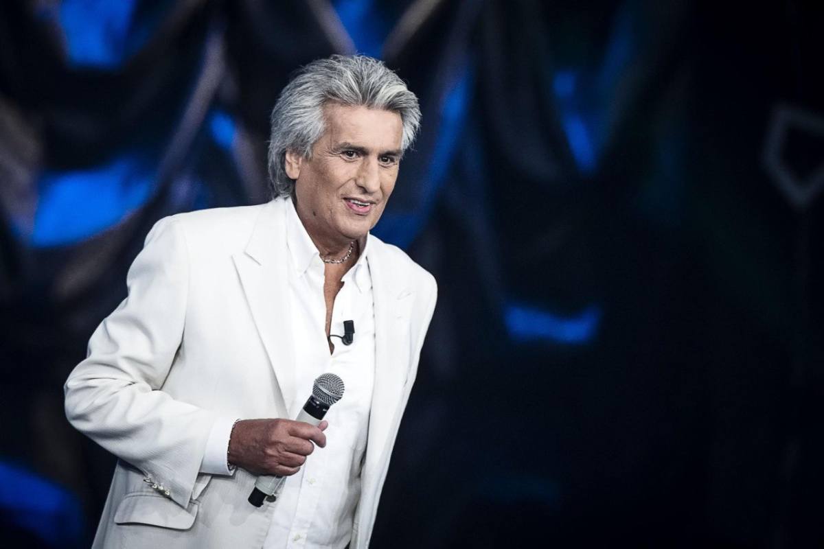 Toto Cutugno da grande in abito bianco e capelli bianchi che tiene un microfono in mano